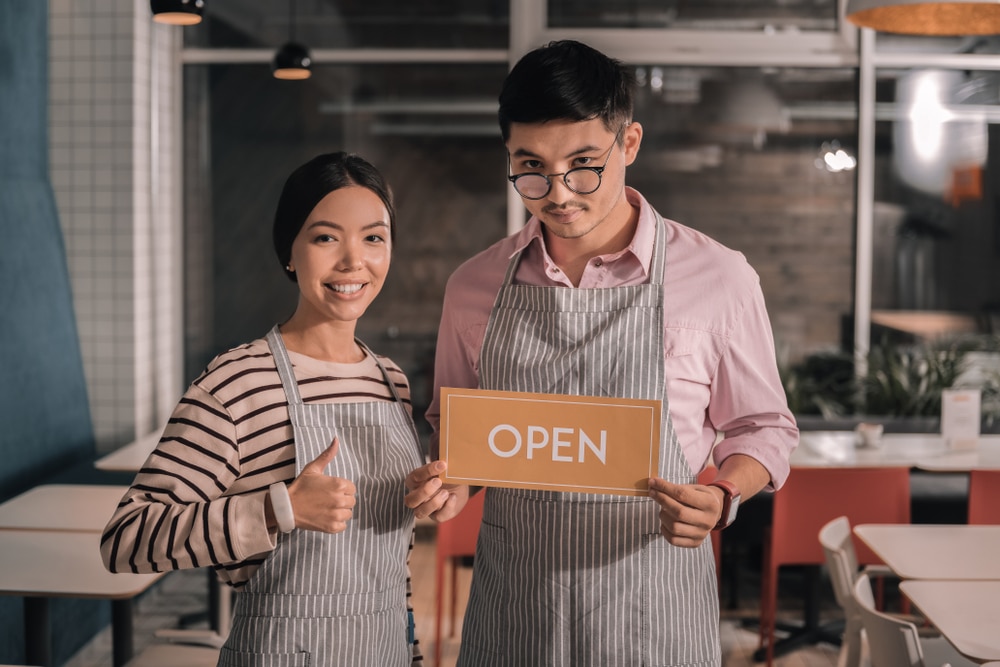 Asian-man-and-woman-holding-Open-sign-inside-cafe