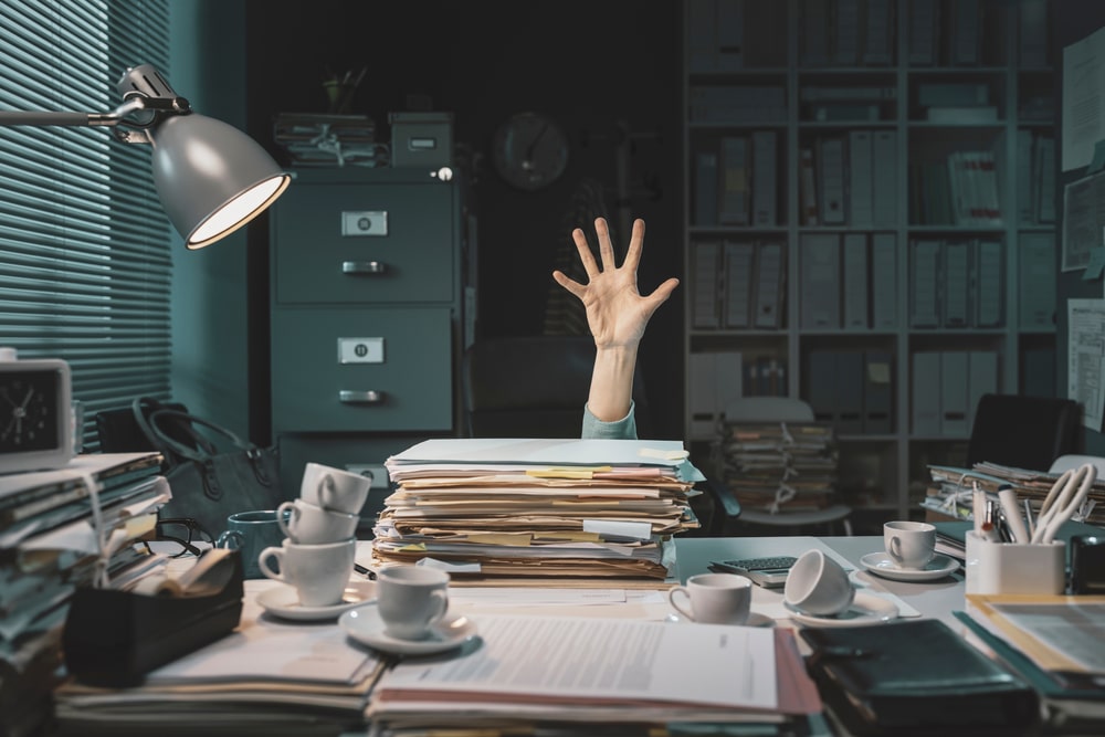 Office-worker-surrounded-by-paperwork-holding-a-hand-in-the-air-from-behind-stack-of-files